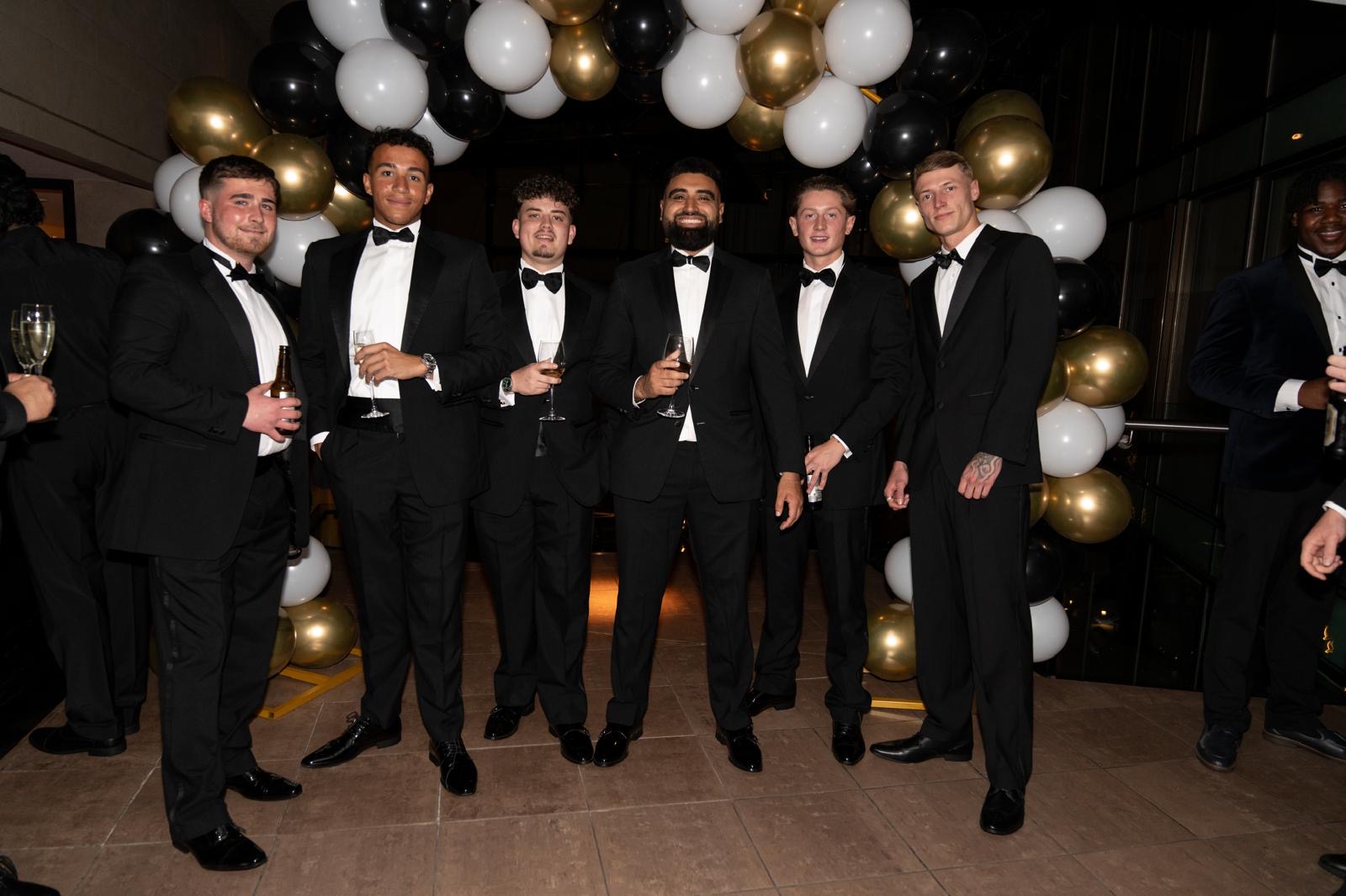 The Elysium team in black tie at the annual agency ball, raising a toast under a black and gold balloon arch.